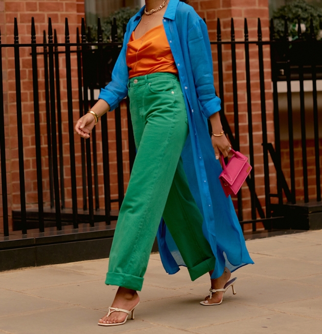 woman in green jeans orange top and blu shirt walking down the street in white sandal